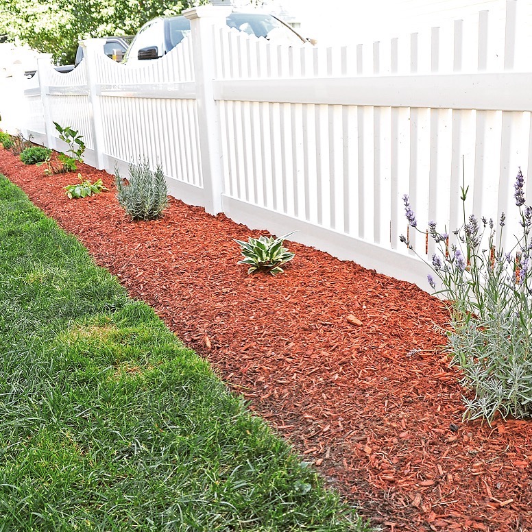 A white picket with mulch and flowers in front of it.