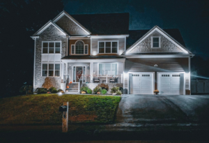 A two-story house with an attached two-car garage is illuminated at night. The home has a stone and siding exterior, a covered front porch, and a mailbox at the edge of a well-maintained lawn.