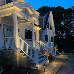 A white, two-story house with a well-lit porch and stone foundation is shown in the evening, surrounded by trees and lit pathway lights leading to the entrance.