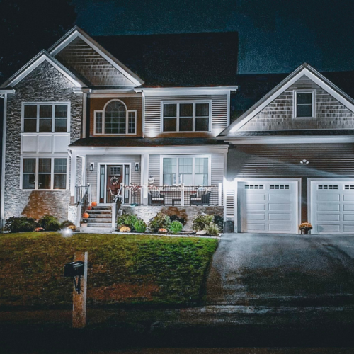 A two-story house with an attached two-car garage is illuminated at night. The home has a stone and siding exterior, a covered front porch, and a mailbox at the edge of a well-maintained lawn.
