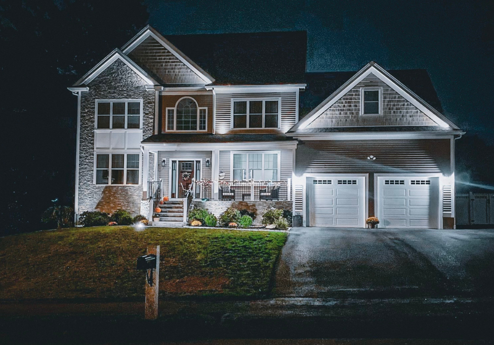 A two-story house with an attached two-car garage is illuminated at night. The home has a stone and siding exterior, a covered front porch, and a mailbox at the edge of a well-maintained lawn.