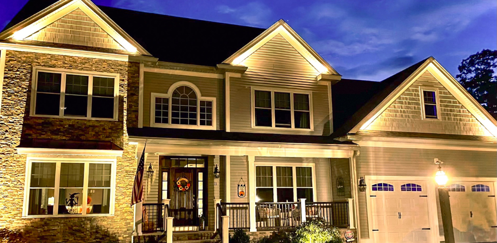 A two-story house with illuminated exterior lights at dusk. It features a stone facade, large windows, and a front porch with decorations. A two-car garage is on the right, and a lawn surrounds the house. Landscape Lighting.