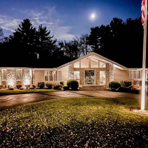 A single-story house with large windows is illuminated at night. The front lawn is neatly manicured, and a tall flagpole with an American flag stands in the yard. A full moon is visible in the night sky.
